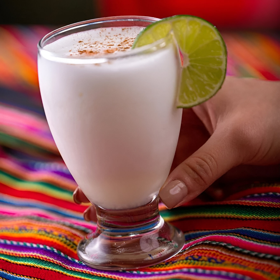Peruvian Pisco Sour cocktail, garnished with a lime slice and cinnamon, held over a colorful textile.