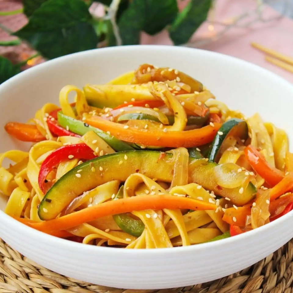 Peruvian Lomo Saltado with thick noodles, colorful vegetables, and sesame seeds in a white bowl.